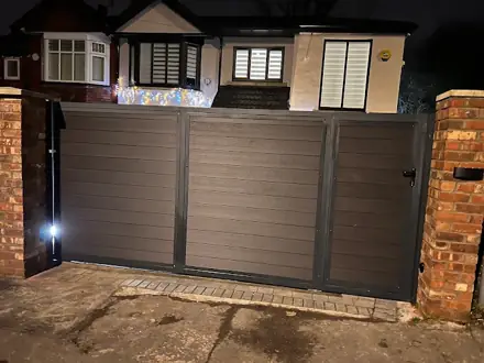 Grey horizontal-board driveway gates between brick pillars, lit after dark