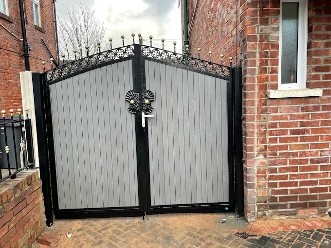 Grey composite double gates in a black frame with arched scrollwork and gold finials, beside a red-brick house